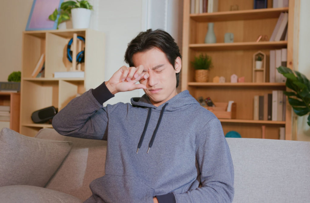 A young man rubbing his right eye with his right hand in the living room at home while sitting on a sofa.