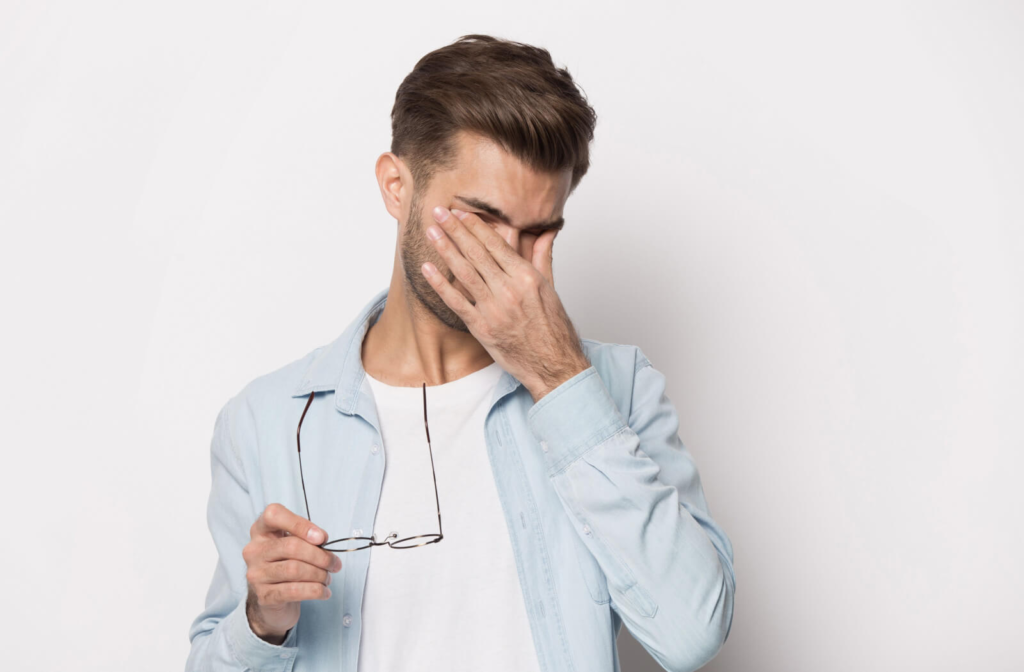 A young man standing and holding his glasses with his right hand as he rubs his eyes with his left hand.
