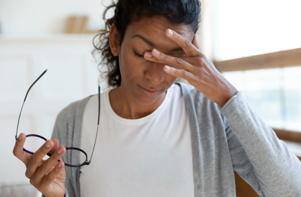 A woman removes her glasses to rub her eyes with her left hand.