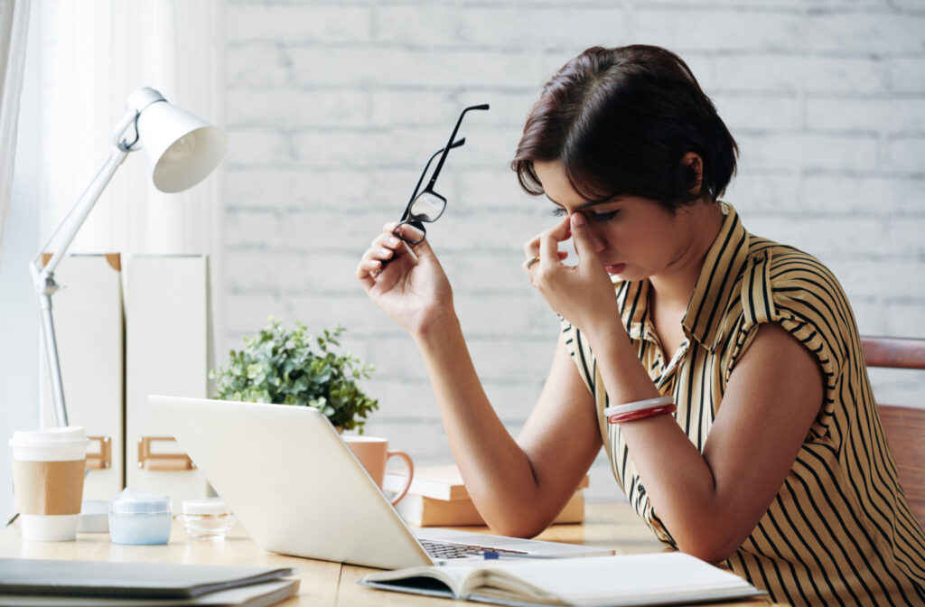 A woman sitting at a desk with a laptop and holding her glasses in her right hand as she holds the bridge of her nose with her other hand.