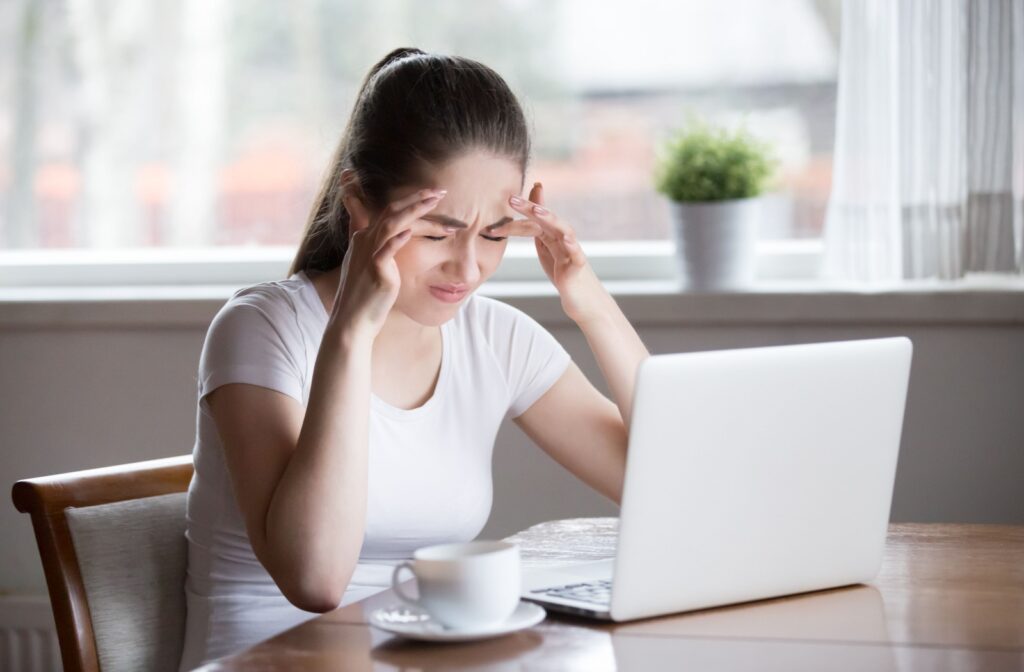 A person sits at a table with a laptop and cup of coffee, holding their head in discomfort from eye strain.
